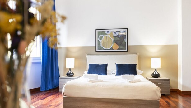 A neatly made bed with white linens and two blue pillows, flanked by two bedside tables with lamps, in a room with wooden flooring and a framed artwork above the bed. - Easylife - Milan - italia 11 P4 - Historic Center (Milano)
