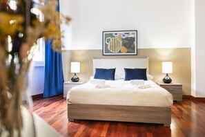 Een net opgemaakt bed met witte lakens en twee blauwe kussens, met aan beide zijden nachtkastjes met lampen, in een kamer met houten vloer en een kunstwerk boven het bed.
