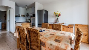 A dining area with a table set for six, surrounded by woven chairs, a kitchen in the background, and a vase with flowers on the table. - Charming apartment with pool, near center and lake, sleeps 4 (Sanguinet)