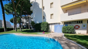 Una piscina con agua cristalina azul, rodeada por un camino empedrado y contigua a un edificio de varios pisos con balcones.