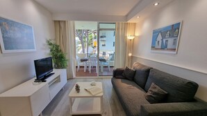 Un salón moderno con un televisor de pantalla plana, una mesa de centro blanca, un sofá gris y un balcón con vistas a edificios y árboles.