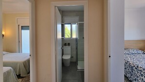 Ein schmaler Flur führt zu einem Badezimmer mit Toilette und Fenster. Auf beiden Seiten des Flurs befinden sich jeweils zwei Betten mit gemusterten Bettbezügen.