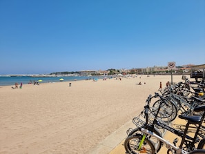 Uma praia com bicicletas estacionadas na lateral, céu limpo e placa indicando 'proibido cães'.