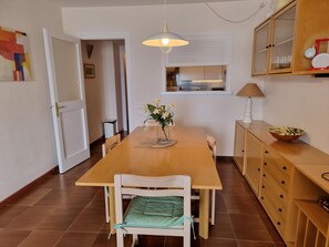 A dining area with a wooden table, chairs, a vase with flowers, a hanging light fixture, and a kitchen area visible through an open door. - Cozy apartment near the beach with air conditioning (L'Escala)