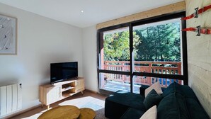 A modern living room with a large window, a television, a wooden cabinet, a green sofa, and a small round table. - Renovated apartment for 6 near slopes - Les 2 Alpes (Les Deux Alpes)