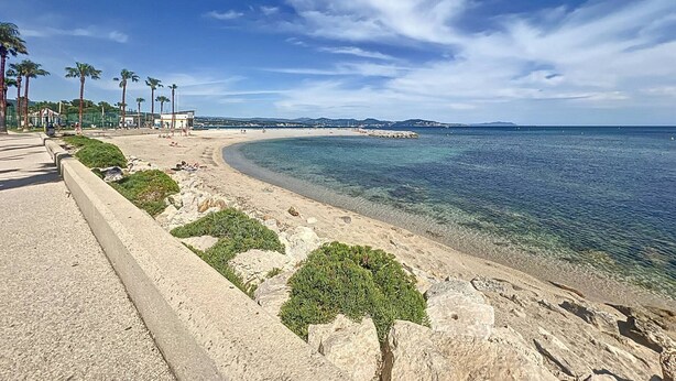 En kustvy med en strand, kristallklart blått vatten, en sandstrand och en strandpromenad som kantas av palmer.