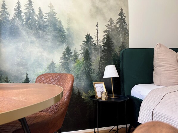Ein Zimmer mit einem Bett, einem kleinen Tisch mit einer Lampe, einem Stuhl und einem Wandbild von einem Wald.