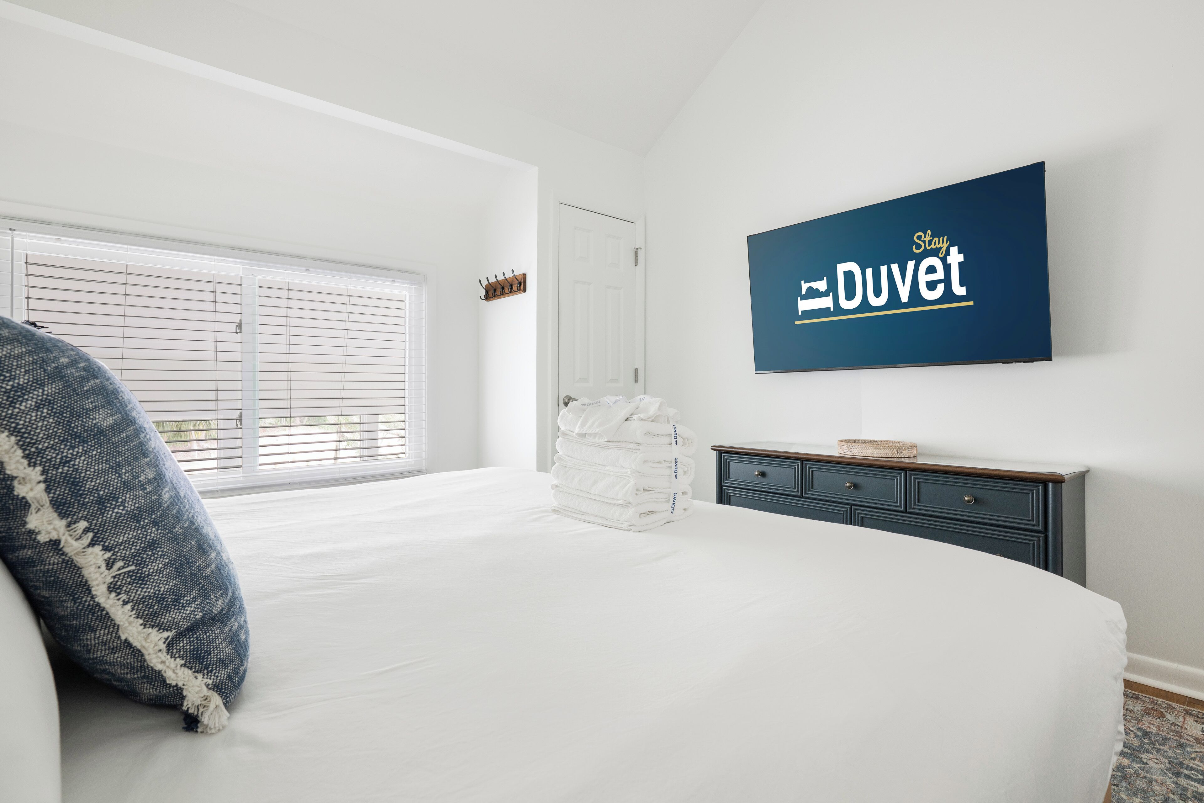 Une chambre avec un grand lit, une fenêtre avec des stores, un téléviseur fixé au mur et une commode sombre.