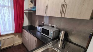 Een kleine keuken met een magnetron, broodrooster en waterkoker op het aanrecht, houten kastjes en een raam met rode gordijnen.