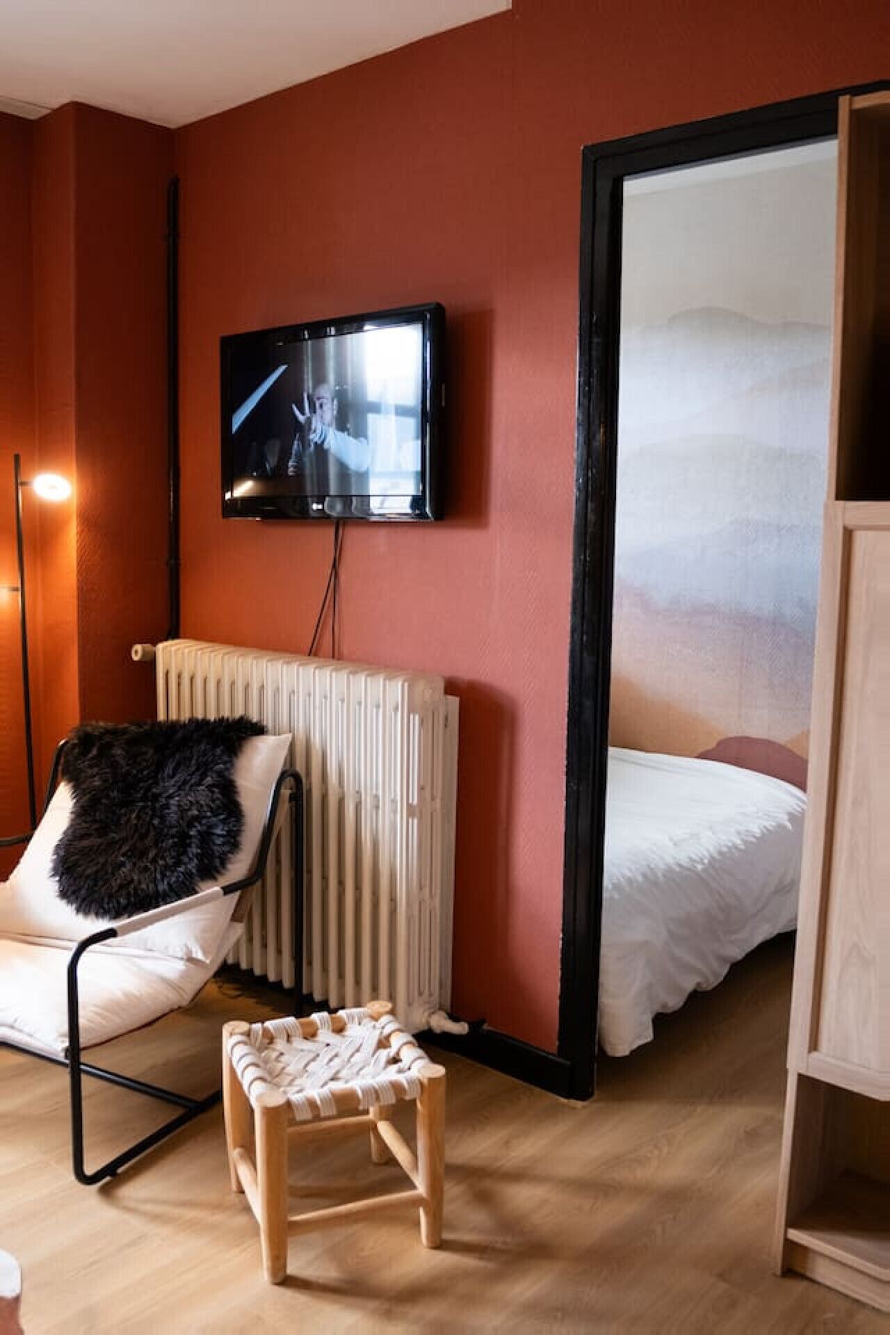 Una habitación con cama, una silla, una mesita pequeña, un radiador y un televisor montado en la pared.