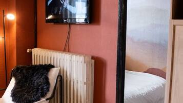 Una habitación con cama, una silla, una mesita pequeña, un radiador y un televisor montado en la pared.