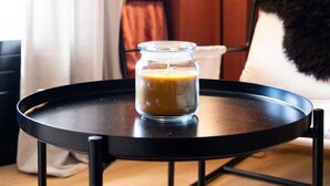 Een glazen pot koffie op een klein rond tafeltje met metalen poten, een zwarte vloerlamp, een witte stoel met een zwart-wit kussen, en een bruin tapijt.