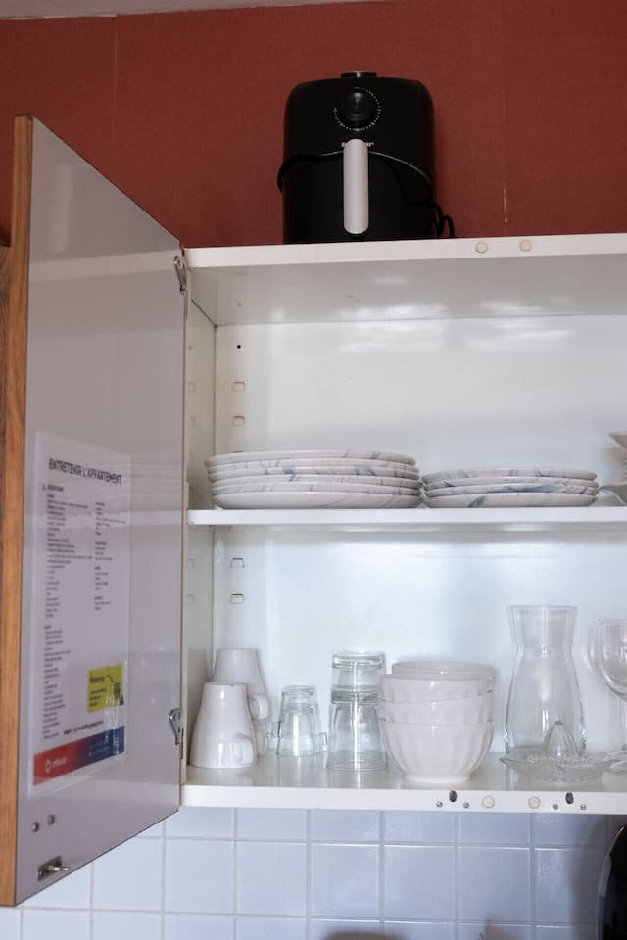 Keittiössä on valkoiset kaapit, joissa on päällekkäin aseteltuja lautasia, sekä vedenkeitin ja lasiesineitä.