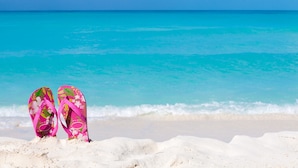 Et par lyserøde flip-flops på en sandstrand med turkis vand.