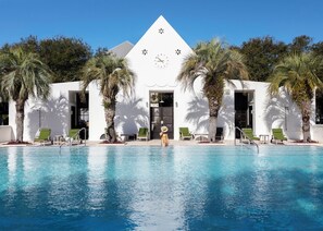 Un bâtiment moderne avec une piscine à l’avant, agrémenté de palmiers et de fauteuils de jardin verts.