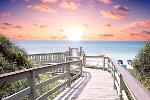 Ein hölzerner Steg führt zu einem Strand, im Hintergrund ist ein Sonnenuntergang zu sehen.