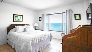 Une chambre à coucher avec un grand lit, une commode en bois, une table de chevet avec une lampe et une fenêtre donnant sur la mer.
