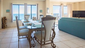 Un espace repas comprenant une table et des chaises, un canapé, un téléviseur et de grandes fenêtres offrant une vue sur l’océan.