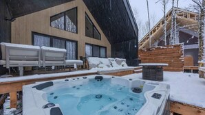 A hot tub in a snowy outdoor area with a wooden deck and a modern building in the background. - Kos - Domaine du Grand Portneuf - Spa, Nature & Co (Pont-Rouge)