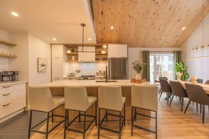 A modern kitchen with a central island, bar seating, and a dining area with chairs. - Kos - Domaine du Grand Portneuf - Spa, Nature & Co (Pont-Rouge)