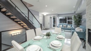 A modern dining area with a glass table set for six, surrounded by white chairs. A staircase with glass railings leads to an upper level. - 👑Luxury Inner City New Built /Garage /AC/EV Charger (Calgary)