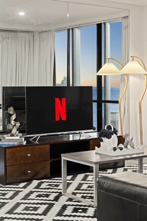 Un saló modern amb un televisor de pantalla plana gran, un moble de roba de nit de fusta, una taula de centre i una vista de la ciutat a través de grans finestres.