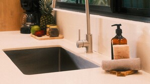 Een keukenkraan met modern design, een houten snijplank met fruit en een koffiezetapparaat, en een raam met uitzicht op groen.