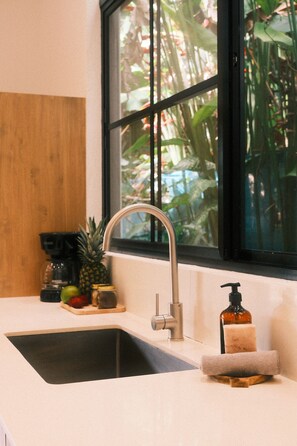 A kitchen sink with a modern faucet, a wooden cutting board with fruits and a coffee maker, and a window with a view of greenery. - Raw. Tropical. Unique. Villa Madom by NP Marino Ballena (Osa)