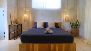 Een slaapkamer met een groot bed, een houten hoofdeinde, twee bedlampen, een kamerplant en een raam met dunne gordijnen.