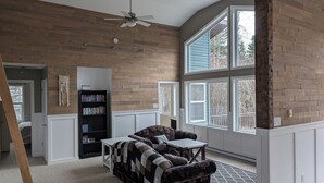 A spacious living room with a large window, a ceiling fan, a bookshelf, and a patterned sofa.