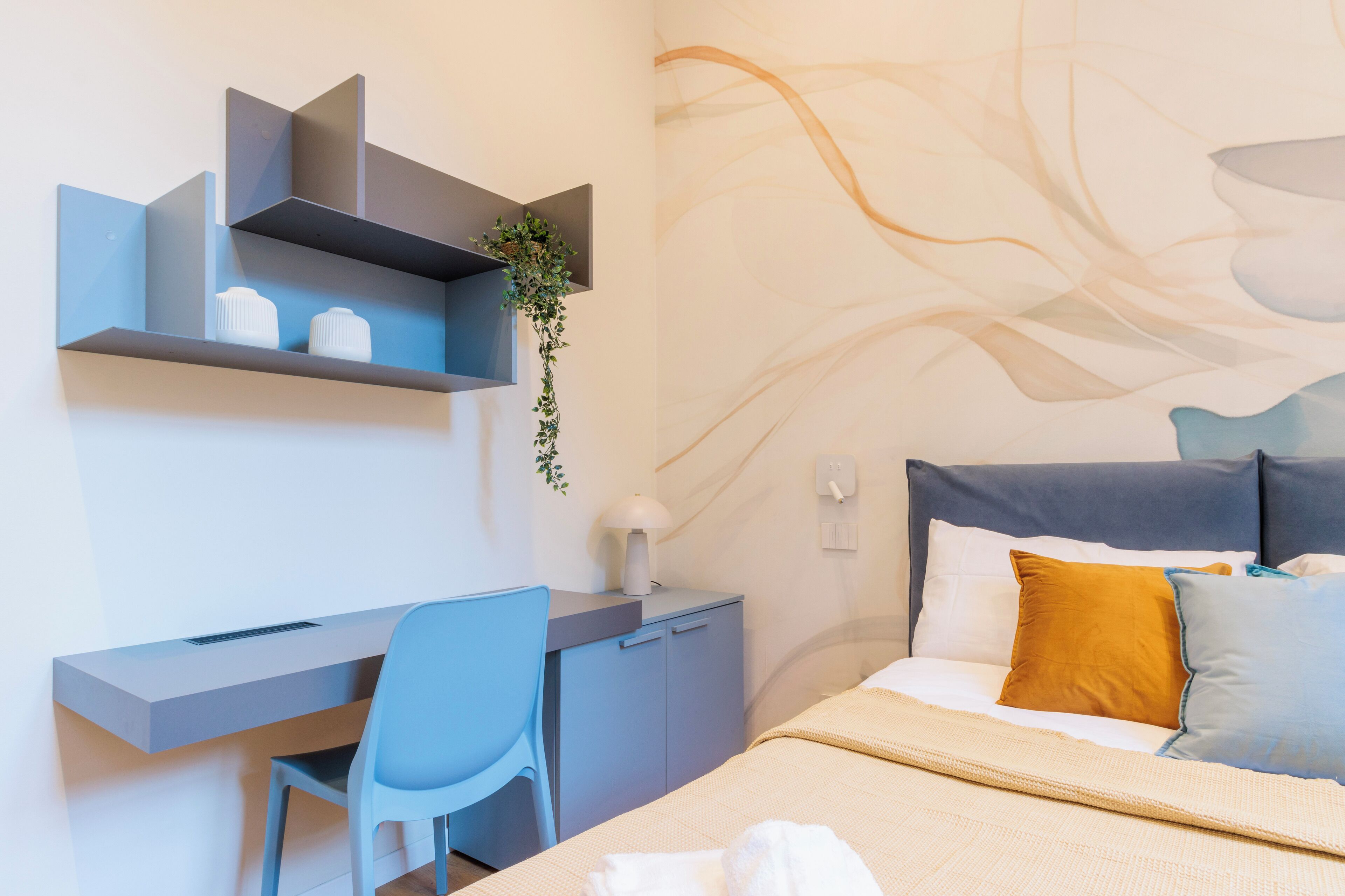 A modern hotel room with a bed, a desk, and a chair. There is a wall-mounted shelf with decorative items and a plant.