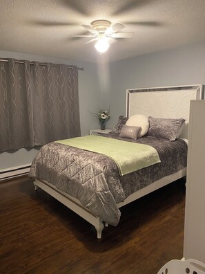 Ein Schlafzimmer mit einem Bett, einem Nachttisch, einer Vase mit Blumen, einem Deckenventilator und einem Fenster mit Vorhängen.