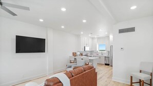 Ein modernes Wohnzimmer mit einer braunen Ledersofa, einem flachbildfernseher an der Wand und einer Küchenzeile mit weißen Schränken und Edelstahlgeräten.