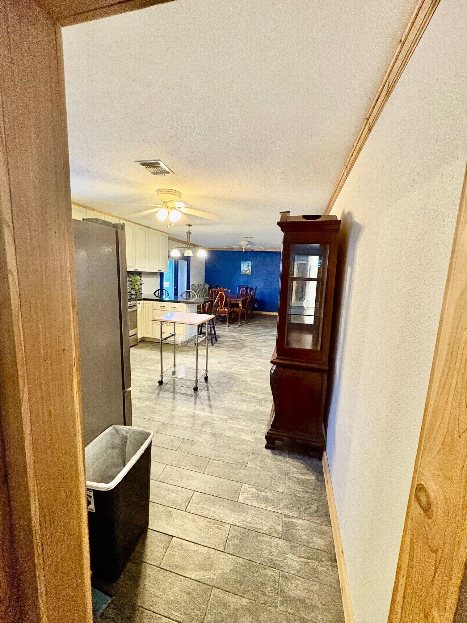 A hallway leading to a kitchen with a dining area, a trash bin, and a wooden cabinet.