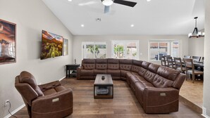 Ein geräumiges Wohnzimmer mit Ecksofa, Liegesessel und Couchtisch. Es gibt einen Deckenventilator, einen Essbereich mit Stühlen und ein Fenster mit Jalousien.