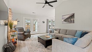 Un salon spacieux avec un grand canapé, une table basse et un ventilateur de plafond. Une porte donne accès à un balcon.
