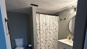 Une salle de bain avec des toilettes, un rideau de douche orné de motifs floraux, un lavabo avec un miroir et un tapis marron sur lequel est inscrit le mot « Naked ».