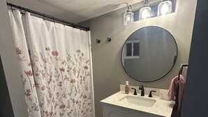 Une salle de bain avec un meuble-lavabo blanc, un miroir rond, un lavabo et un rideau de douche orné de motifs floraux.