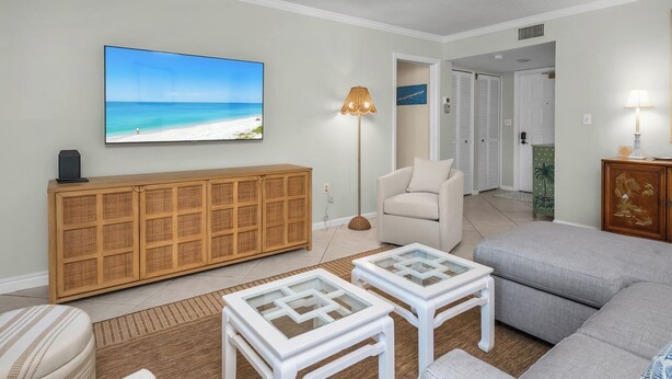 Una sala de estar con un televisor de pantalla plana grande, un mueble de consola de mimbre, un sillón color beige y un sofá gris.