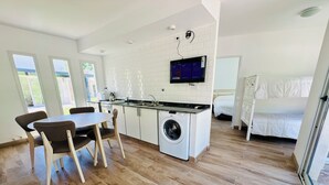 Une cuisine moderne équipée d’un lave-linge intégré, un coin repas avec des chaises en bois et un lit dans la pièce adjacente.