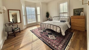 A bedroom with a large window, a bed with a patterned bedspread, a wooden dresser, and a mirror. - Hip Historical First Fl 1BR Downtown King size bed (Brunswick)