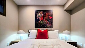 Una cama bien hecha con sábanas blancas y una almohada roja, dos lámparas de noche y un cuadro de una bicicleta en la pared.