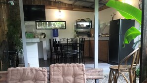 Un espacio al aire libre acogedor con bancos acolchados y una mesa de comedor, rodeado por una terraza de madera rústica y una estructura de techo de paja.