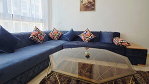 Una sala de estar moderna con un sofá seccional azul, almohadones estampados y coloridos, una mesa de centro de vidrio con base geométrica y un cuadro en la pared.