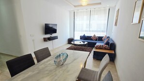 Una sala de estar moderna con comedor, un conjunto de sofás, televisor montado en la pared y un gran ventanal con cortinas.