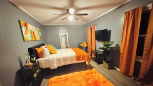 Ein Schlafzimmer mit einem Bett, Deckenventilator, Fernseher, Vorhängen und einer Pflanze.