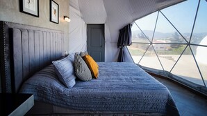 A bedroom with a large bed, a headboard, a door, and a window with a view of the landscape. - Cupulas Del Valle  (Valle de Guadalupe)