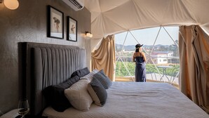 A tented accommodation room with a bed, pillows, a framed picture, an air conditioning unit, and a view of the landscape. - Cupulas Del Valle  (Valle de Guadalupe)