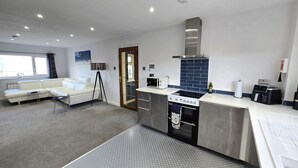 A modern kitchen with a built-in stove, microwave, and coffee maker. There is a white sofa and a floor lamp in the adjacent living area. - Spacious apartment, 2 mins walk to the beach! (Perranporth)