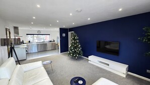 A modern living room with a white sofa, a Christmas tree, a flat-screen TV, and a kitchen in the background. - Spacious apartment, 2 mins walk to the beach! (Perranporth)